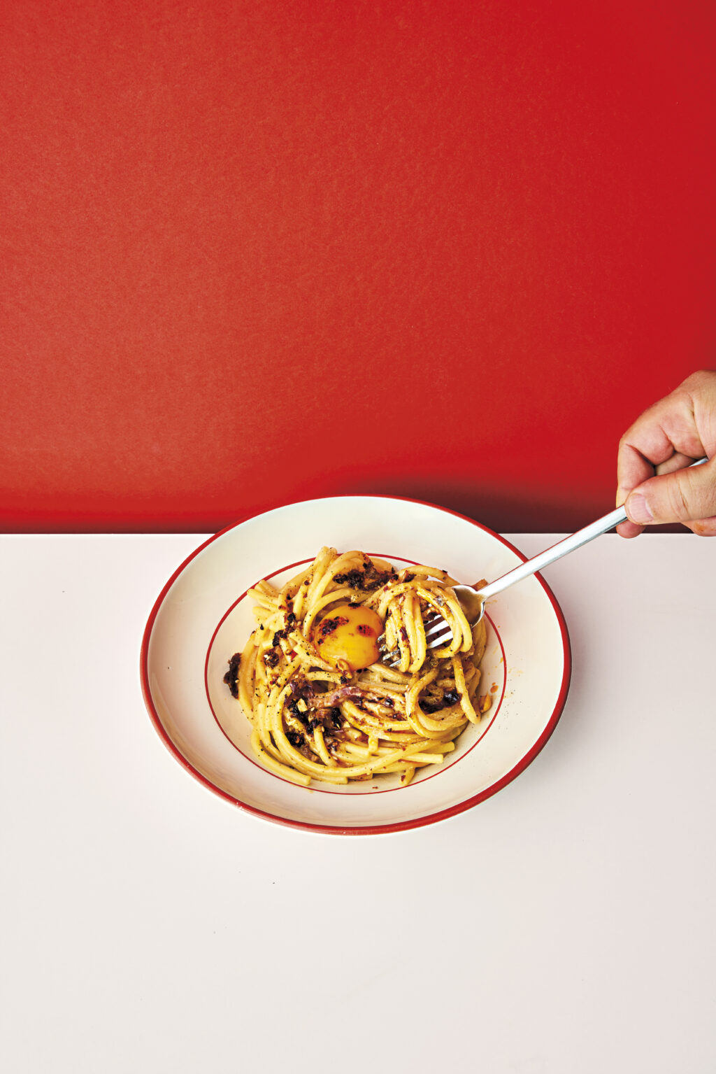 En tallrik carbonara med chilicrisp. Vitt bord med röd vägg i bakgrunden.