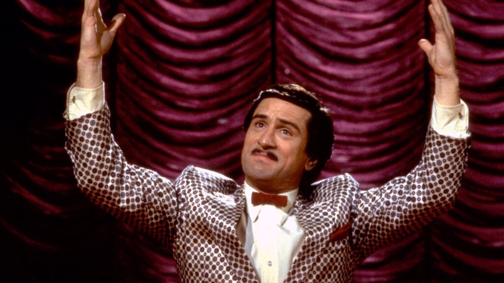 Robert De Niro i filmen King of Comedy. Han har kostym, står framför ett draperi och höjer armarna uppåt.