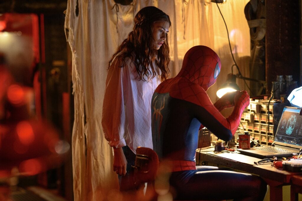 MJ tittar över axeln på Spider-Man som sitter vid ett arbetsbord framför en dator.