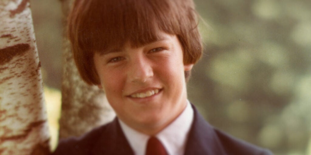 "Smiling boy with brown hair in a suit and tie, standing outdoors next to a birch tree, blurred green natural background."